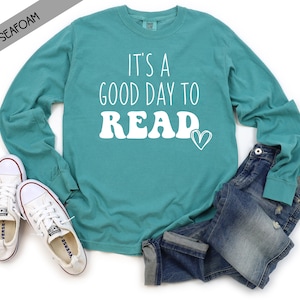 May include: A teal long-sleeve t-shirt with the text "It's a good day to READ" in white with a heart design. The shirt is shown on a white background with a pair of white sneakers and blue jeans.