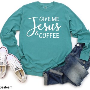 May include: A teal long-sleeve t-shirt with the text "Give Me Jesus & Coffee" in white lettering. The shirt is shown on a white background with a pair of white sneakers and blue jeans.