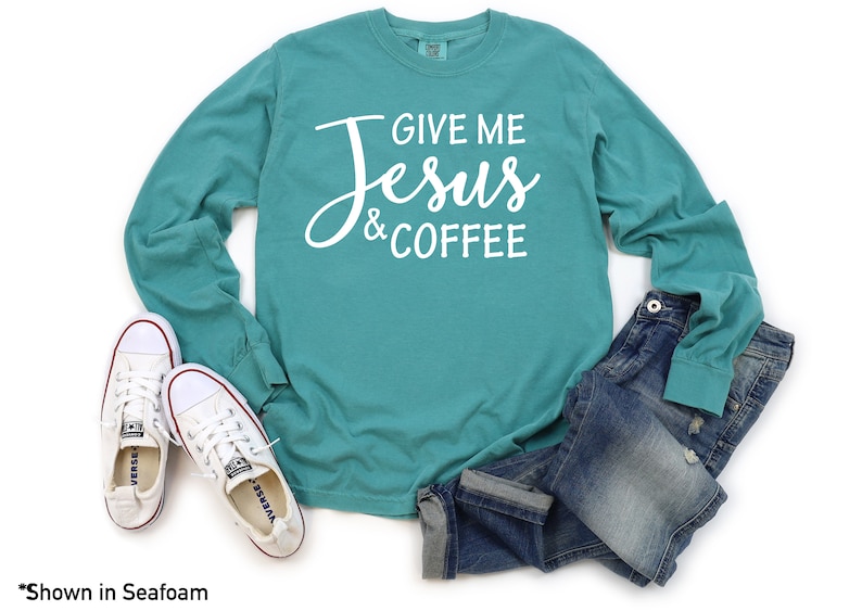 May include: A teal long-sleeve t-shirt with the text "Give Me Jesus & Coffee" in white lettering. The shirt is shown on a white background with a pair of white sneakers and blue jeans.