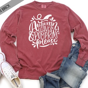 May include: A long-sleeved t-shirt in a brick red color with the text "Autumn Leaves & Pumpkins Please" in white lettering.