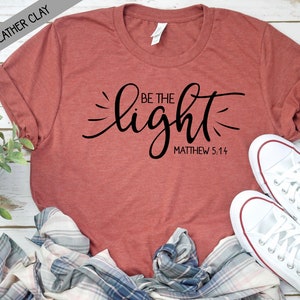 Puede incluir: Una camiseta de color óxido con el texto "Be the light" en letras cursivas negras y "Matthew 5:14" en texto negro debajo. La camiseta está sobre una superficie de madera con una camisa a cuadros y zapatillas blancas.