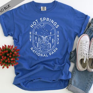 May include: A bright blue short-sleeve t-shirt featuring a white graphic print of a landscape with a waterfall, trees, and stars inside an oval frame. The text reads HOT SPRINGS NATIONAL PARK EST 1832. The shirt is paired with blue jeans.
