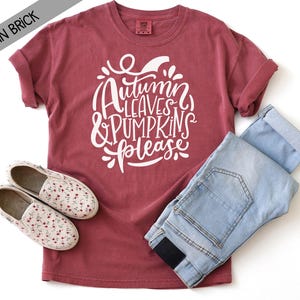 May include: A brick red short-sleeve t-shirt featuring white cursive text that reads Autumn Leaves & Pumpkins Please, paired with light blue denim jeans and a pair of white slip-on shoes with a small red floral pattern throughout the fabric.