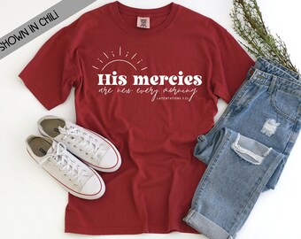 May include: A chili red short-sleeve t-shirt featuring a white graphic of a sun and the text "His mercies are new every morning" with "LAMENTATIONS 3:23" below it. The shirt is paired with white canvas sneakers and distressed blue denim jeans.