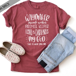 May include: A brick red short-sleeve t-shirt featuring white text that reads: WAYMAKER miracle worker PROMISE KEEPER LIGHT IN THE DARKNESS MY GOD THAT IS WHO YOU ARE. The shirt is paired with light blue denim jeans and white floral slip-on shoes.