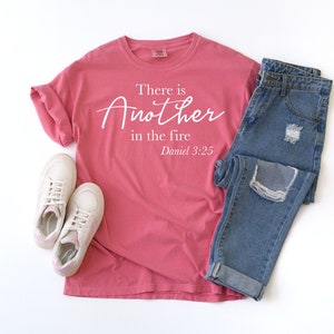 May include: A pink t-shirt with the text "There is Another in the fire Daniel 3:25" in white lettering. The shirt is paired with blue jeans and white sneakers.