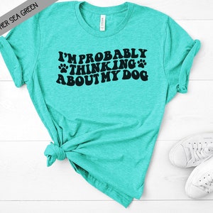 May include: A turquoise blue t-shirt with the text "I'm probably thinking about my dog" in black lettering with paw prints.