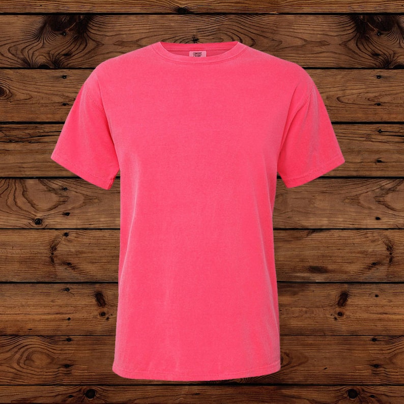 May include: A solid coral-colored short-sleeve t-shirt. The shirt has a crew neck and a slightly faded appearance. The fabric appears to be soft and comfortable, suitable for casual wear. The background is a wooden surface.
