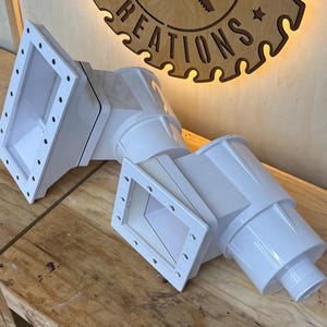 May include: A white, angled structure with rectangular openings, possibly for ventilation or ductwork. It rests on a wooden surface, with a wooden sign in the background that reads "Chad's Custom Creations, Est. 2012."