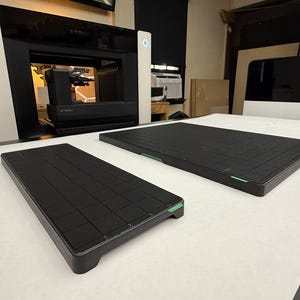 May include: Two rectangular black build plates with a grid pattern surface and small green accents on the edges. These flat, solid components are positioned on a white surface in front of a large, white and black 3D printer with an X logo on top.