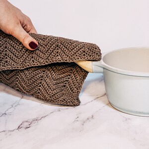 May include: A brown knitted pot holder with a wooden handle, being placed on a white marble surface next to a white ceramic pot.