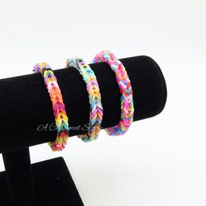 May include: Three colorful rubber band bracelets displayed on a black velvet stand. The bracelets are woven in a fishtail pattern, featuring a mix of vibrant colors including pink, yellow, orange, blue, and green. The bracelets are approximately 0.75 inches wide.