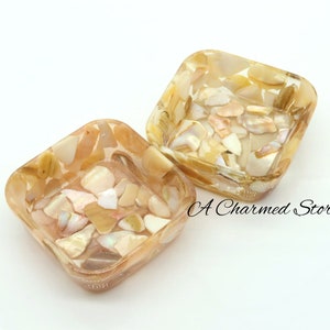 May include: Two square-shaped decorative bowls made of clear resin with embedded iridescent shell pieces. The bowls are a light beige color with a subtle shimmer.