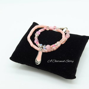 May include: A pink and silver bracelet with a pink ballet shoe charm. The bracelet is made of pink beads and silver beads. The charm is attached to the bracelet with a silver ring. The bracelet is on a black velvet cushion. The text "A Charmed Story" is visible on the cushion.