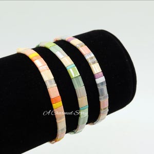 May include: Four colorful beaded bracelets displayed on a black velvet surface. Each bracelet features a different color scheme, including orange, yellow, pink, green, and purple. The bracelets are made of small, rectangular beads.