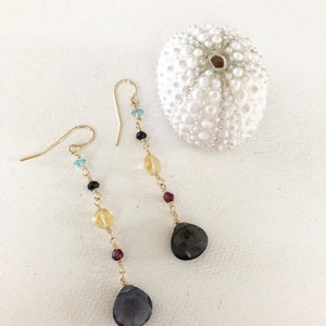 May include: A pair of long, gold dangle earrings with a teardrop-shaped gemstone at the bottom. The earrings feature a variety of colorful beads, including blue, black, yellow, and red.