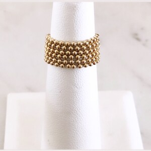 Op de afbeelding: Een ring van goudkleurige, kleine ronde kralen. De ring is gestapeld met meerdere rijen kralen, waardoor een textuur en geometrisch ontwerp ontstaat.