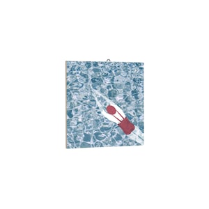 May include: A square art print depicts a swimmer in a red swimsuit diving into a pool of blue water. The water's surface has a rippled effect, and the swimmer is seen from above, creating a serene and summery scene.