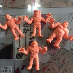 Peut inclure: Cinq petites figurines d'action de couleur pêche dans diverses poses sont présentées sur fond d'illustration de bande dessinée. Les figurines semblent être des personnages de lutte ou de combat, avec des corps musclés détaillés.