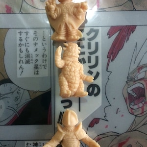 Peut inclure: Trois petites figurines en plastique beige de personnages de la série d'anime Dragon Ball Z. Les figurines sont posées dans différentes poses d'action. Le texte "クリリンの" est visible en arrière-plan.