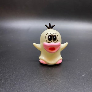 May include: A white ceramic figurine of a cartoon ghost with black hair, pink cheeks, and a wide open mouth. The ghost has large black eyes and is standing on a black surface.