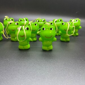 May include: A group of 10 green frog figurines with white eyes and a string attached to each. The frogs are all facing forward and have a smiling expression. The frogs are all the same size and shape.