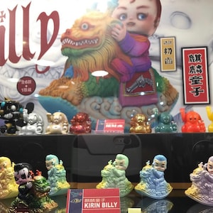 May include: A display case featuring a collection of Kirin Billy figurines. The figures are in various colors, including gold, red, and blue. The main figure is a baby riding a colorful lion-like creature. The text "KIRIN BILLY" is visible.