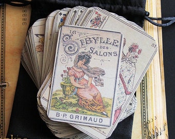 Könnte beinhalten: Ein Deck Vintage-Spielkarten in einem schwarzen Samtbeutel. Die Karten zeigen eine Frau in einem rosa Kleid und einem weißen Schal mit dem Text "La Sibylle des Salons B.P. Grimaud" auf der Vorderseite.