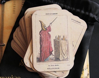 May include: A deck of vintage playing cards with a worn, aged look. The cards are stacked in a pile and feature a black and white illustration of a person in a red robe holding a staff. The text on the card reads "Le faux devin. MELANCOLIE."