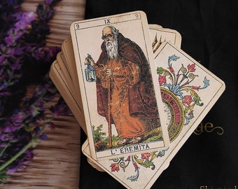 May include: A vintage tarot deck with the Hermit card face up. The card features a man with a long white beard and a lantern, walking with a staff. The card is surrounded by ornate floral designs.