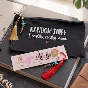 May include: A black zippered pouch with the white text "RANDOM STUFF I really, really need." and a small red heart, featuring a gold tassel and turquoise starfish charm. A rectangular bookmark with a pink flamingo, "Aloha" text, and a red tassel.