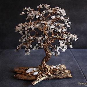 Könnte beinhalten: Eine handgefertigte dekorative Baumskulptur mit einem gedrehten Kupferdrahtstamm und Ästen. Die Äste sind mit kleinen, klaren und grauen Edelsteinen geschmückt, die Blättern ähneln. Der Baum ist auf einem Stück Naturholz montiert.