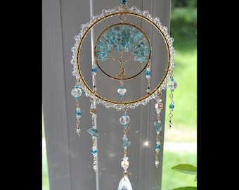 May include: A decorative hanging ornament featuring a gold-toned wire hoop wrapped in clear beads. Inside, a smaller gold wire tree sculpture with light blue stone leaves is suspended. Multiple dangling chains with blue and clear beads hang below.