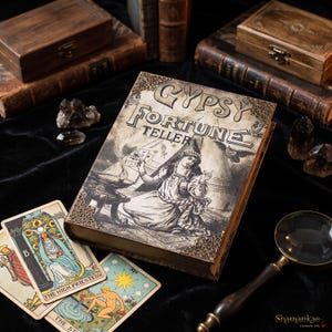 Può includere: Un libro intitolato Gypsy Fortune Teller con un'illustrazione in stile vintage di una persona che tiene in mano delle carte, caratterizzato da angoli in metallo dorato. Il libro è circondato da due carte dei tarocchi, La Papessa e La Stella, una lente d'ingrandimento e cristalli scuri.