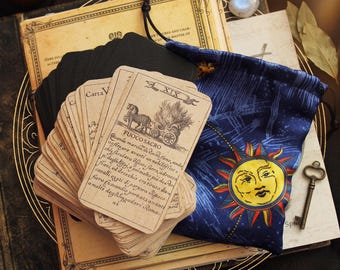 May include: A deck of beige tarot cards with black ink illustrations and Italian text, including a card labeled XIX Fuoco Sacro. The deck is paired with a dark blue drawstring pouch featuring a yellow sun graphic and a small metal skeleton key.