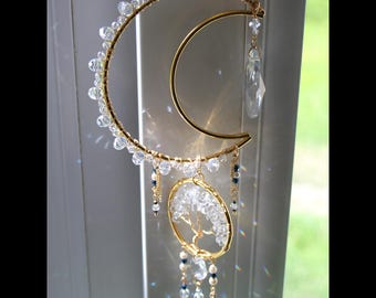 May include: A gold-colored metal suncatcher featuring a crescent moon and a circular tree of life design. The moon is adorned with clear crystal beads. Additional clear crystals and blue beads dangle from the design. The suncatcher hangs from a gold chain.