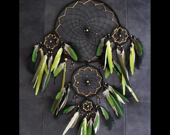 May include: A black dreamcatcher with gold accents and green feathers. The dreamcatcher has three hoops, with the largest hoop being the outermost. The dreamcatcher is decorated with black beads and gold beads.