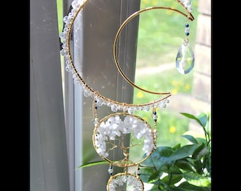 May include: A gold-colored metal and crystal suncatcher with a crescent moon and two circular frames. The frames feature a tree design and are filled with white stones. Clear crystals and a teardrop-shaped crystal dangle from the suncatcher.