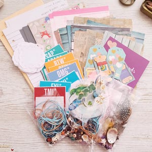 May include: A collection of stationery supplies including lined paper, patterned cardstock, and small tags with text like TMI, OMG, BTW, and ASAP. The set includes clear plastic bags filled with blue and orange twine, wooden beads, and small feathers.
