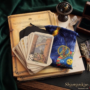 Può includere: Un mazzo di carte dei tarocchi con una carta che mostra due animali e un arcobaleno, appoggiato su una pila di documenti in stile vintage. Accanto alle carte c'è un sacchetto blu con coulisse con illustrazioni celesti e una piccola sfera armillare.
