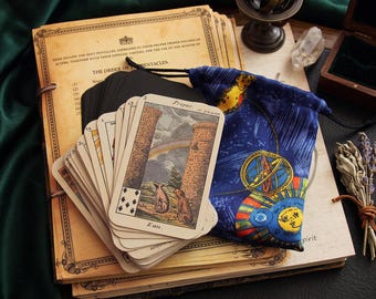 May include: A deck of tarot cards with a card showing two animals and a rainbow, resting on a stack of vintage-style paper documents. Beside the cards is a blue drawstring pouch featuring celestial illustrations and a small brass armillary sphere.