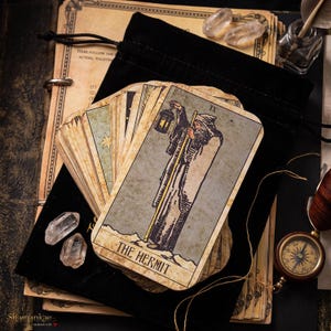 Può includere: Un mazzo di carte dei tarocchi con un'illustrazione in stile vintage dell'Eremita sulla carta superiore, che mostra una figura barbuta che regge una lanterna e un bastone. Il mazzo è appoggiato su un sacchetto di velluto nero con due quarzi.