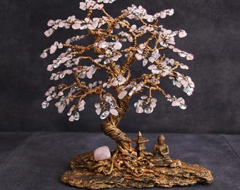 May include: A handcrafted wire tree sculpture with a bronze-colored trunk and branches, adorned with pink and clear crystal gemstones. A small Buddha statue and a pink gemstone are at the base. The tree is on a textured, bronze-colored base.