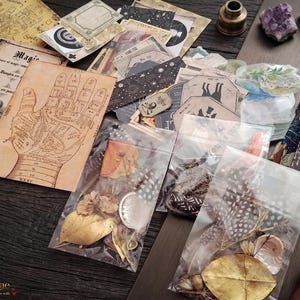 Peut inclure: Une collection d'éphémères comprenant des cartes en papier de style vintage avec des diagrammes de chiromancie, des illustrations célestes et occultes, et des sachets en plastique transparent contenant des éléments naturels comme des feuilles dorées, des plumes tachetées et des petits coquillages.