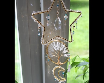 May include: A hanging suncatcher featuring a gold wire star frame, a circular tree of life design, and a crescent moon. The piece is adorned with clear crystal beads, small blue accents, and a central teardrop-shaped crystal prism hanging from the star.