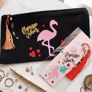 Könnte beinhalten: Ein schwarzes Reißverschlussetui mit einem rosa Flamingo-Motiv, roten Herzen und dem goldenen Schriftzug "flamingo vibes", dazu ein passendes Lesezeichen in einer Klarsichthülle, ein goldener Füllfederhalter, Metallringe und ein Kompass.
