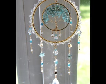 May include: A decorative hanging ornament featuring a gold-toned wire hoop adorned with clear beads and a central tree of life motif crafted from gold wire and small blue-green stone chips. Multiple beaded strands with blue accents hang below.