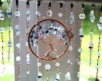 May include: A copper-toned, wire-wrapped tree of life wall hanging with a circular frame, filled with blue, purple, and clear stones. Strands of beads and crystals hang below, creating a decorative, bohemian-style window or wall art piece.