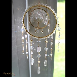 Puede incluir: Un atrapasoles decorativo con dos aros de alambre concéntricos en tono dorado. El aro interior contiene una escultura de árbol hecha de alambre con hojas de cristal transparente. El aro exterior está adornado con cuentas, cadenas y colgantes.