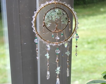 May include: A circular hanging ornament featuring a gold-toned wire tree with light green stone chips as leaves, centered within a larger gold hoop. The outer hoop is adorned with clear beads, while dangling strands feature clear crystals and green stones.